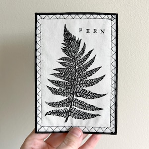 May include: A black and white embroidered patch featuring a fern design and the word "FERN" in black text.