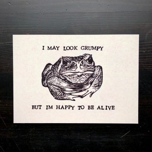 May include: A black and white illustration of a grumpy-looking toad with the text "I may look grumpy but I'm happy to be alive".