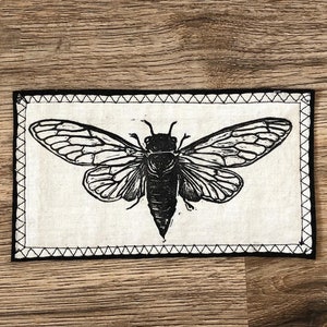 May include: Black and white illustration of a cicada insect printed on a white fabric patch with a black stitched border.