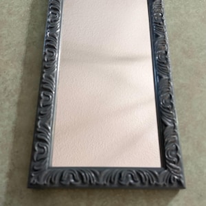 May include: A rectangular wall mirror featuring a dark gray frame with a raised, repeating scrollwork pattern. The reflective glass surface is clear and smooth, set within the ornate, textured border that surrounds all four sides.