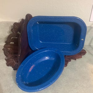 May include: A rectangular blue enamel baking pan and an oval blue enamel bowl, both featuring a white speckled pattern. The rectangular pan has a small hole on one side, and the oval bowl has a small hole on its rim, both resting against a dark brown carved wooden decorative piece.