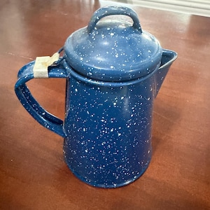 May include: A blue enamel coffee percolator with a white speckled pattern throughout the body and lid. The percolator features a curved handle with a metallic silver-colored attachment at the top and a spout on the opposite side of the handle.