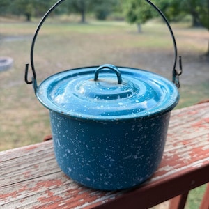May include: A teal enamel pot with a white speckled pattern, featuring a matching lid with a small handle and a thin, dark metal wire bail handle attached to the sides. The pot has a cylindrical shape and a smooth, glossy finish throughout.