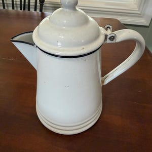May include: A white enamel coffee pot with a matching lid, a curved handle, and a spout. The pot features a thin black rim around the top edge and the lid, with a small knob on the center of the lid. The body has a smooth finish with subtle details.