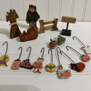 May include: A collection of wooden Halloween-themed ornaments and figurines. The set includes a scarecrow, a fence, a sign reading Pumpkins 25 cents, a corn shock, pumpkins, and various hanging ornaments featuring a witch, ghost, bat, owl, and cat.