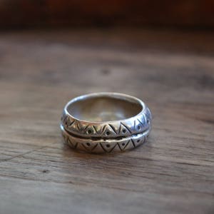 May include: A silver ring with a geometric pattern. The ring is made of two bands, each with a different pattern. The ring is on a wooden surface.