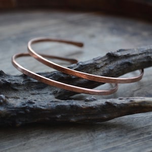 Peut inclure: Deux bracelets en cuivre empilés l'un sur l'autre, reposant sur un morceau de bois usé.
