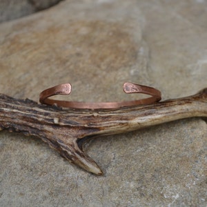 May include: A simple copper bracelet with a hammered finish. The bracelet is open ended and sits on a piece of antler.