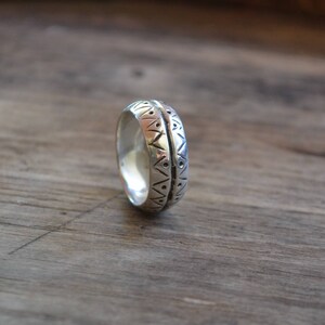 May include: A silver band ring with a geometric pattern engraved around the entire band.