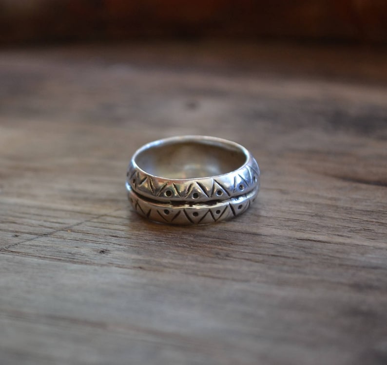 May include: A silver ring with a geometric pattern. The ring is made of two bands, each with a different pattern. The ring is on a wooden surface.