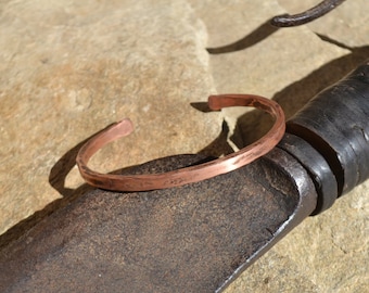Può includere: Un bracciale a polsino in rame con una texture martellata. Il bracciale aperto è di un caldo colore marrone-rossastro e sembra fatto a mano. Il bracciale è appoggiato su una superficie marrone strutturata.