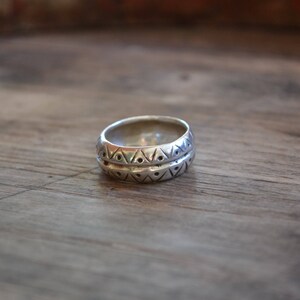 May include: A silver ring with a geometric pattern. The ring is wide and has a textured surface.