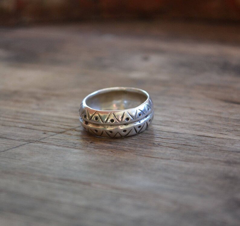 May include: A silver ring with a geometric pattern. The ring is wide and has a textured surface.