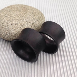 May include: Two black wooden ear gauges, also known as plugs or tunnels, are shown on a textured surface. The gauges are cylindrical with a flared opening at one end.