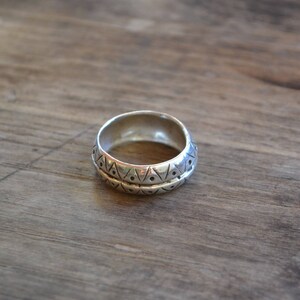 May include: A silver ring with a geometric pattern. The ring is wide and has a rounded shape.