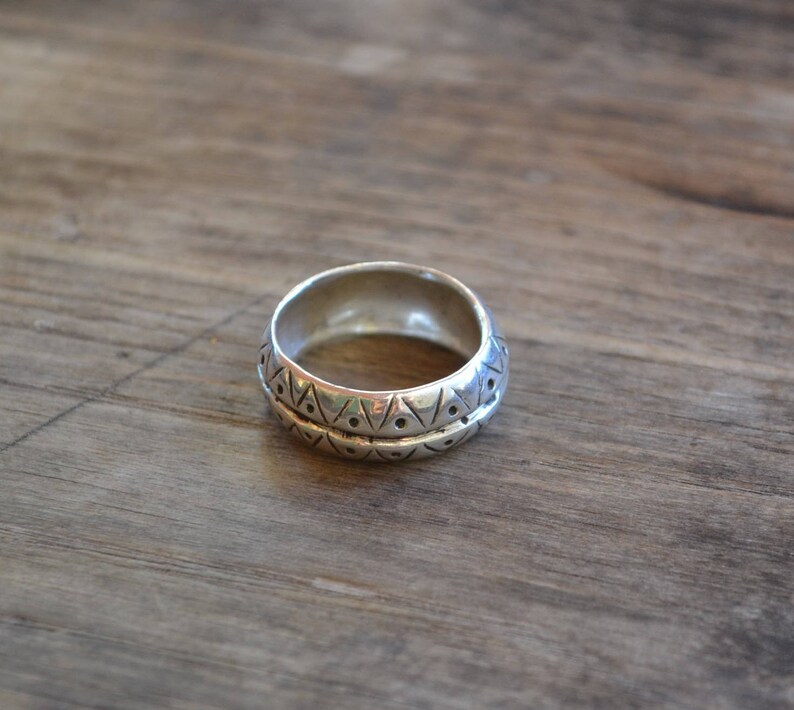 May include: A silver ring with a geometric pattern. The ring is wide and has a rounded shape.