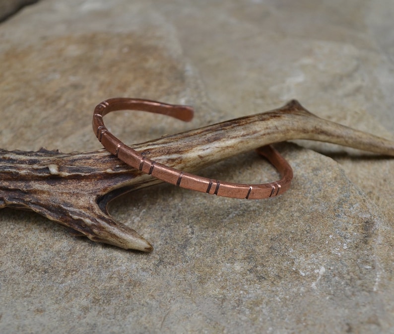 May include: A copper bracelet with a textured design. The bracelet is on a light brown surface.