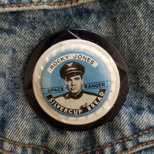 May include: A black and white button with a picture of a man in a uniform. The button says "Rocky Jones Space Ranger Silvercup Bread".