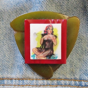 May include: A green and red plastic pin with a picture of a woman in a black dress. The woman is sitting on a yellow background.