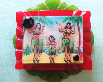 May include: A red, green, and black plastic brooch with a picture of three hula dancers in front of palm trees. The brooch has a scalloped edge and two black rhinestones.