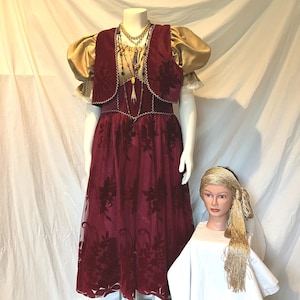 May include: A burgundy velvet dress with a floral pattern, featuring a matching vest and gold puff sleeves. The outfit is accessorized with a layered gold necklace and a gold headscarf with long fringe, displayed on a mannequin and a head form.