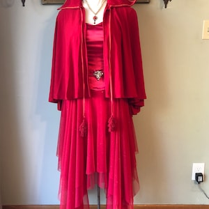 May include: A vibrant red formal dress ensemble. The outfit includes a floor-length dress with a layered tulle skirt, a matching cape with a high collar, and a decorative brooch. A necklace with a red pendant adds a touch of elegance.
