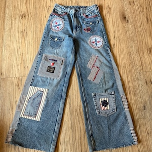 May include: Light blue denim jeans with wide legs, featuring various patches and embellishments. Patches include compass rose designs, striped fabric, and a pocket with a small label. Red stitching accents the details.