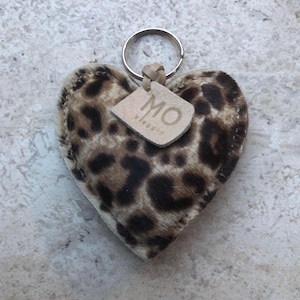 May include: Heart-shaped keychain with a leopard print pattern in shades of brown and tan. The keychain has a metal ring and a small leather tag with the letters "MO" printed on it.