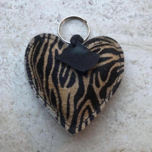 May include: Heart-shaped keychain with a zebra print pattern in black and tan. The keychain has a silver metal ring and a small black leather tag attached. The tag has the word "forever" printed on it.