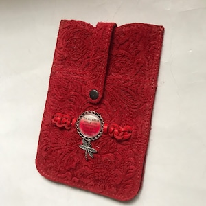 Op de afbeelding: Rode leren telefoonhoes met een bloemreliëfpatroon. De hoes heeft een drukknoopsluiting en een decoratieve bedel met een libellenhanger en de tekst "DA MI BASIA MILLE". Gevlochten rode lederen accenten zijn ook aanwezig.