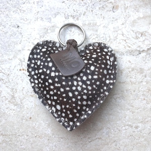 May include: Heart-shaped keychain with a speckled pattern in black and white. The keychain has a silver ring and a small brown leather tag with the letters "MO" printed on it. The background is a light-colored, textured surface.