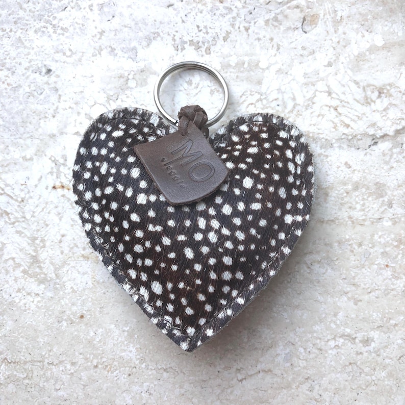 May include: Heart-shaped keychain with a speckled pattern in black and white. The keychain has a silver ring and a small brown leather tag with the letters "MO" printed on it. The background is a light-colored, textured surface.