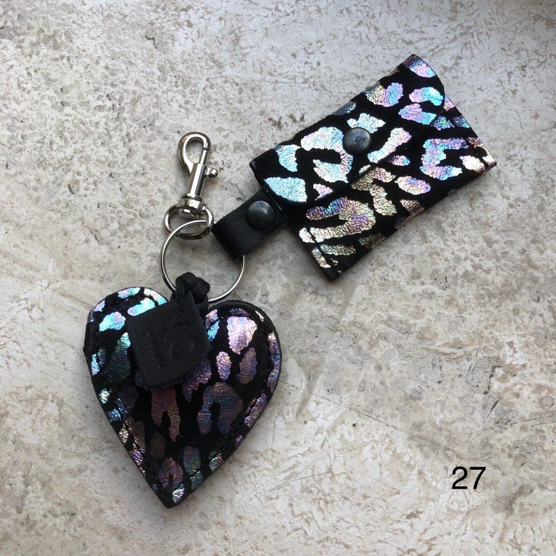 May include: A keychain featuring a heart-shaped charm and a small rectangular pouch, both crafted from black material with a shimmering, iridescent leopard print pattern. The components are connected by a silver-toned metal lobster clasp and ring.