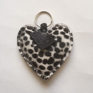 May include: A heart-shaped keychain with a black and white spotted pattern, resembling a Dalmatian's coat. It features a silver key ring and a small, black leather tag.