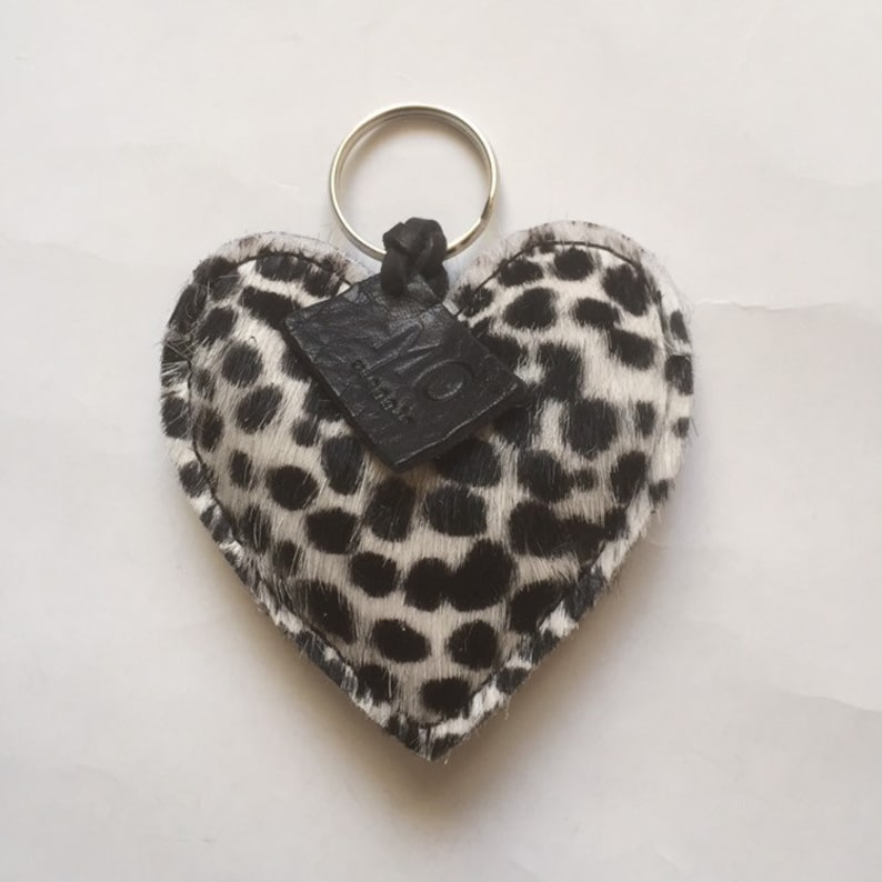 May include: A heart-shaped keychain with a black and white spotted pattern, resembling a Dalmatian's coat. It features a silver key ring and a small, black leather tag.