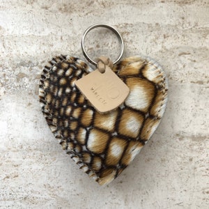 May include: Heart-shaped keychain with a patterned design in shades of brown, tan, and white. The keychain has a metal ring and a small leather tag with the text "MO vicente". The edges are stitched with a contrasting thread.