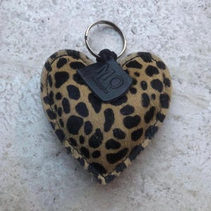 May include: Heart-shaped keychain with a leopard print pattern in brown and black. The keychain has a metal ring and a small black leather tag with the letters "MO" and the word "pioggia" printed on it.