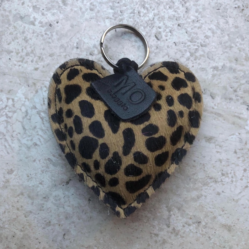 May include: Heart-shaped keychain with a leopard print pattern in brown and black. The keychain has a metal ring and a small black leather tag with the letters "MO" and the word "pioggia" printed on it.