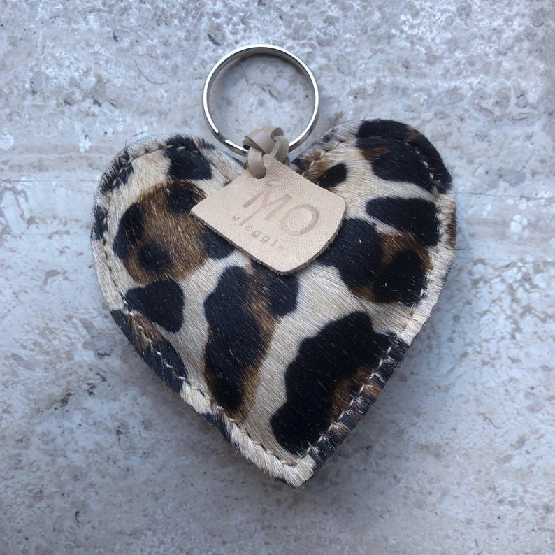 May include: A heart-shaped keychain with a leopard print pattern in shades of brown and cream. The keychain has a silver ring and a small leather tag with the letters "MO" and the word "viaggi" printed on it.