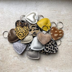 Op de afbeelding: Een verzameling hartvormige sleutelhangers met diverse texturen en patronen, waaronder metallic zilver, felgeel, luipaardprint, cheeta-print, pluizige bruine vacht en gestructureerd grijs leer, elk bevestigd aan een ronde metalen ring.
