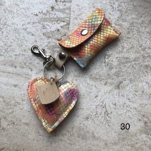 May include: A keychain featuring a small rectangular pouch and a heart-shaped charm, both crafted from a multicolored snakeskin-patterned material. The set includes a silver-tone metal lobster clasp, a split ring, and a small beige leather tag with the embossed text MO. The number 30 is visible in the corner.