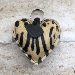 May include: Heart-shaped keychain with a spotted pattern in black and tan. The keychain has a silver ring and a small black leather tag with the letters "MO". The edges are stitched with a contrasting thread.