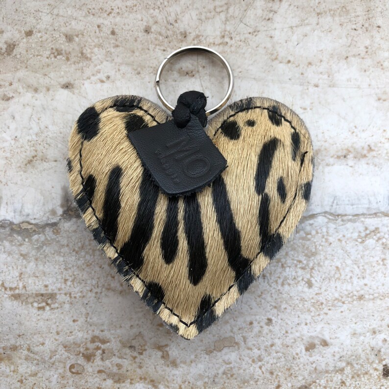 May include: Heart-shaped keychain with a spotted pattern in black and tan. The keychain has a silver ring and a small black leather tag with the letters "MO". The edges are stitched with a contrasting thread.