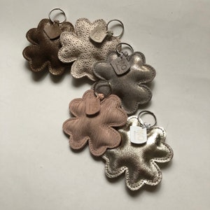 Peut inclure: Plusieurs porte-clés en cuir en forme de trèfle à quatre feuilles. Les couleurs incluent l'or, le marron et le rose. Chaque porte-clés est équipé d'un anneau métallique et d'une petite étiquette avec le texte "MO".