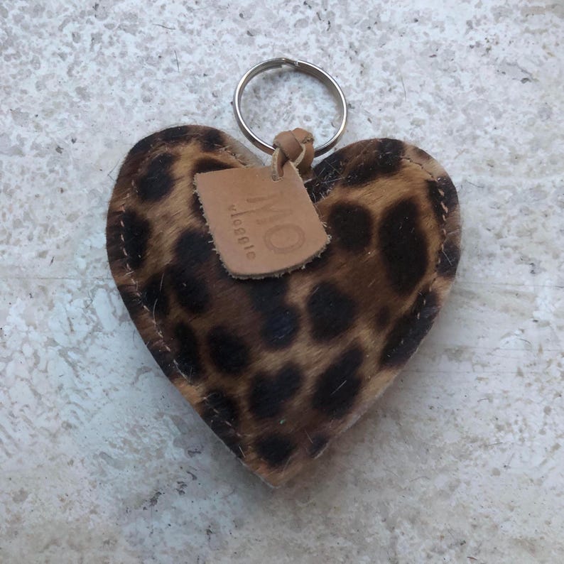 May include: Heart-shaped keychain with a leopard print pattern. The keychain has a metal ring and a small leather tag with the letters "MO" and the word "Maggie" printed on it. The keychain is made of a soft, textured material.
