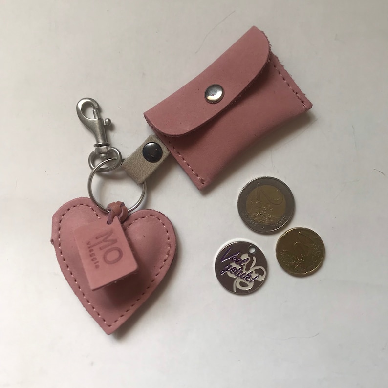 May include: A dusty pink leather keychain featuring a small rectangular pouch with a silver snap button, a heart-shaped charm embossed with the text MO, a metal lobster clasp, and two loose coins, one with the text Veel geluk, on a white surface.