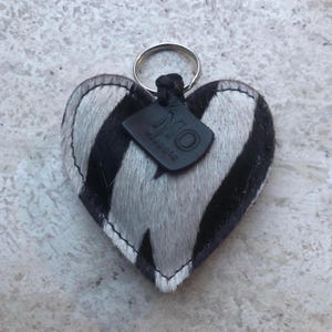 May include: Heart-shaped keychain with a zebra print pattern in black and white. The keychain has a silver ring and a small black leather tag with the letters "MO" and the word "assolo" printed on it.