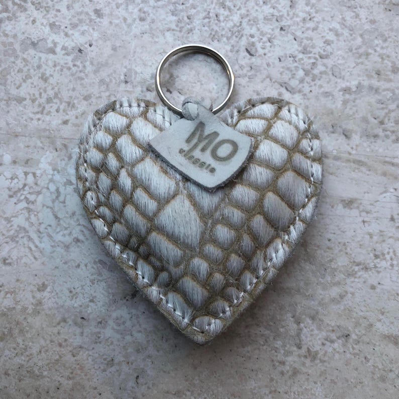 May include: Heart-shaped keychain with a textured, light-colored pattern. The keychain has a silver ring and a small tag with the letters "MO" and the word "classic". The heart is stitched with a contrasting thread.
