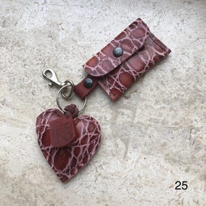 May include: A brown leather keychain featuring a heart-shaped charm and a small rectangular pouch with a snap closure. Both pieces have a textured, embossed pattern. A silver-toned metal lobster clasp and ring connect the items. The number 25 is visible.