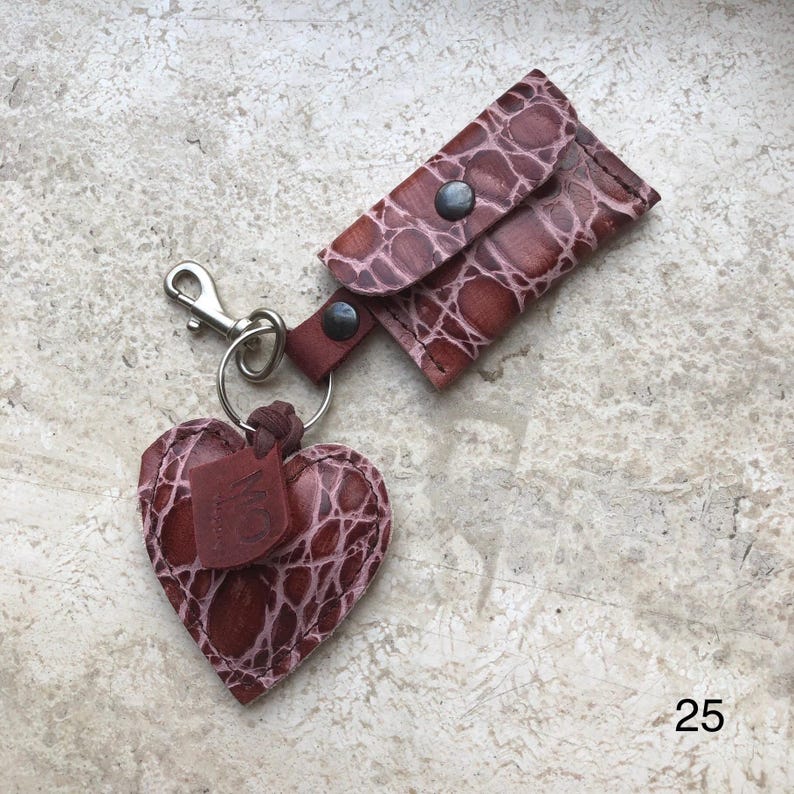May include: A brown leather keychain featuring a heart-shaped charm and a small rectangular pouch with a snap closure. Both pieces have a textured, embossed pattern. A silver-toned metal lobster clasp and ring connect the items. The number 25 is visible.